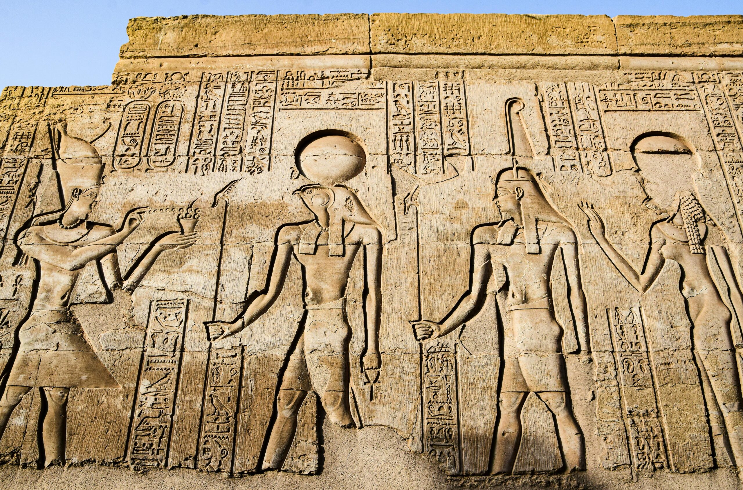 Detailed carvings of ancient Egyptian deities and hieroglyphs on a temple wall in bright daylight.